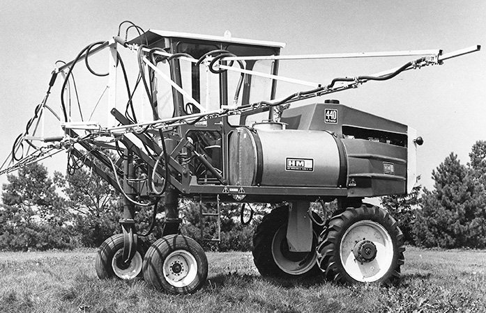 A high-clearance sprayer in a farm field has a cab for the driver and booms sticking out each side.