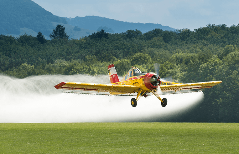 A crop duster flies over a green crop and mists it with pesticide.