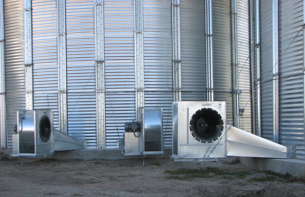 Large fans are attached to the bottom of two large, steel grain bins.