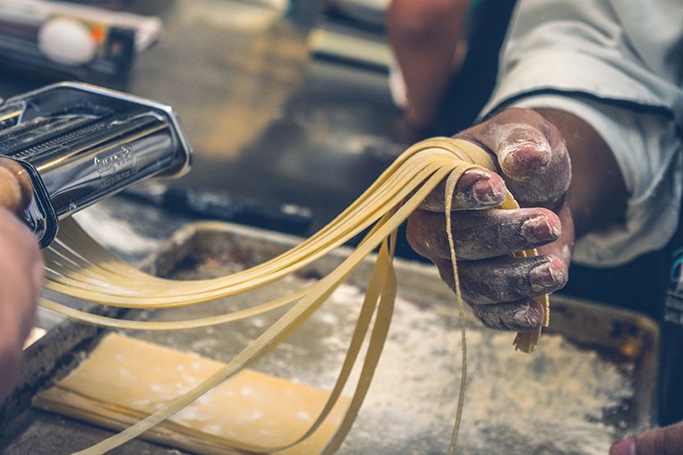 Photograph of pasta being made by putting dough through a hand-held pasta cutter.