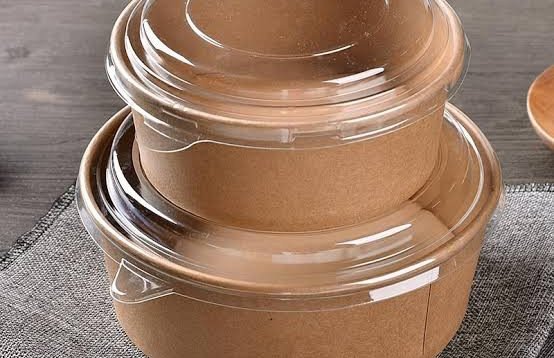 Two paper-looking take-out containers with plastic lids are stacked on top of each other on a wooden table.