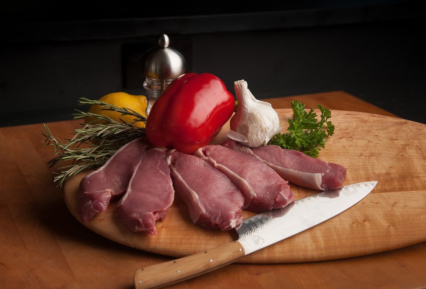 Present-FarmAnimals-Alpaca-Nutrition Five alpaca steaks lie next to each other on a wooden platter next to a pepper, garlic, lemon, and spices.