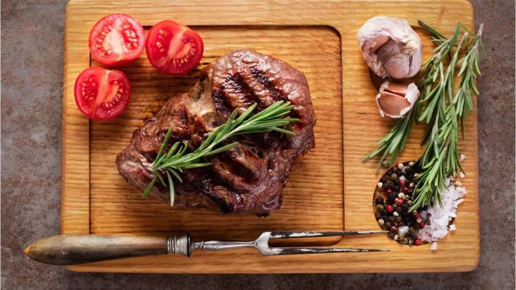 A piece of cooked goat meat sits on a cutting board with tomatoes, salt, pepper, and other herbs.