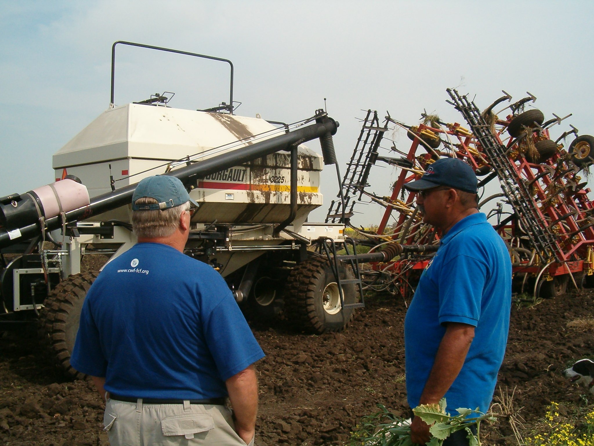 An Indigenous farmer stands talking to a Caucasian person. There is an air-seeder in the background.