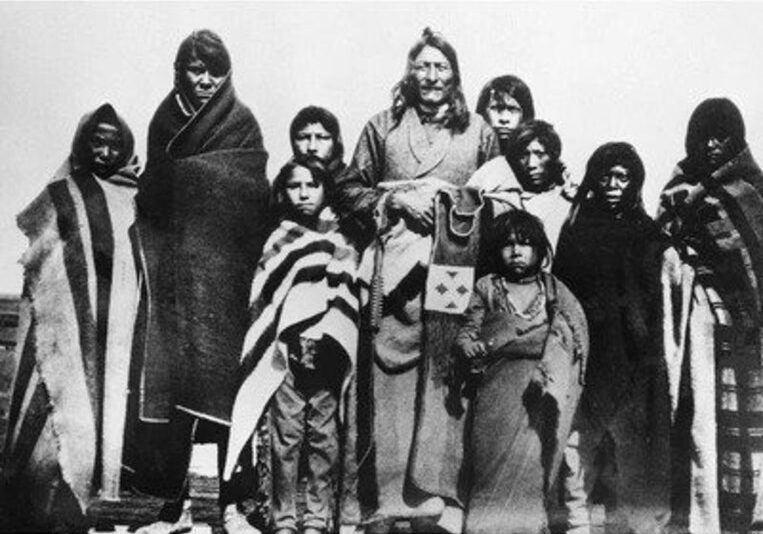 A group of Indigenous people are grouped together, wrapped tightly in blankets. Crowfoot stands in the middle with nine children around him.