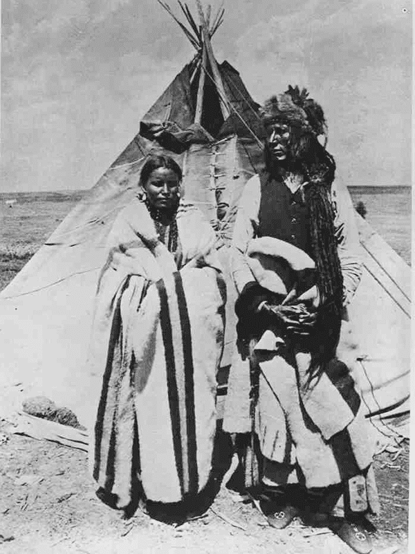 An Indigenous man and woman are wrapped in blankets and stand in front of a tipi.