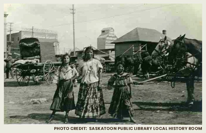 A photograph of three Indigenous girls in skirts posing beside two harnessed horses. A wagon and grain elevator are in the background.