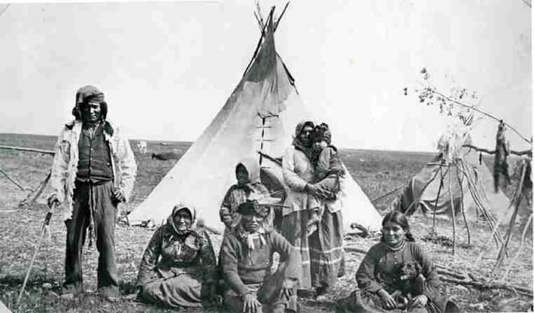 A photograph of Chief Whitecap, Dakota chief of Moose Woods reserve, and members of his family grouped outside two tipis on the plain. The white-haired chief sits on the ground with two women while a younger man, a child, and a woman with a child stand near them.