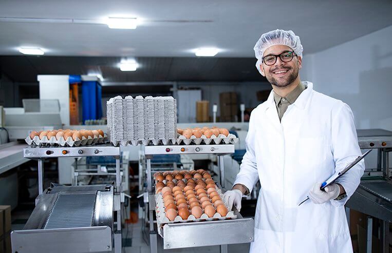 A man stands in a facility next to a line of eggs that are in packages.