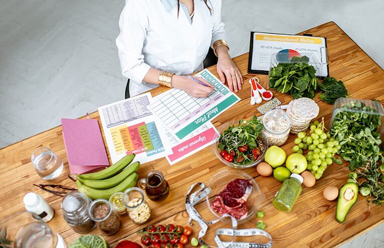 A dietician sits at a table writing on charts. She is surround by vegetables and fruits on the table.