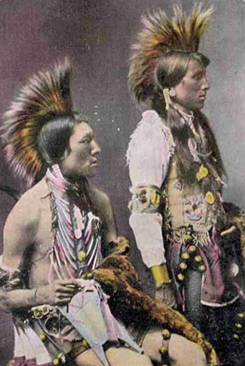 An image of two First Nations men in a side profile studio photograph. They are dressed in ceremonial clothing and each is holding a fur pelt. Colours have been added to the photograph in a chromolithograph process.
