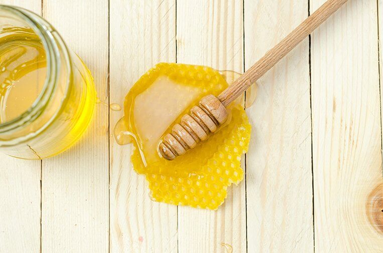 A wooden honey dipper lays on a table with honey on it next to a honey jar.